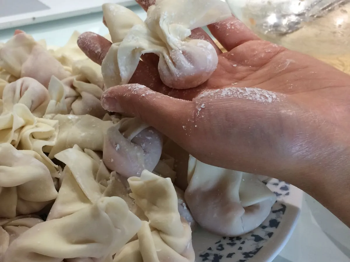 A floury hand holding a freshly wrapped Shanghai-style wonton over a plate of wontons